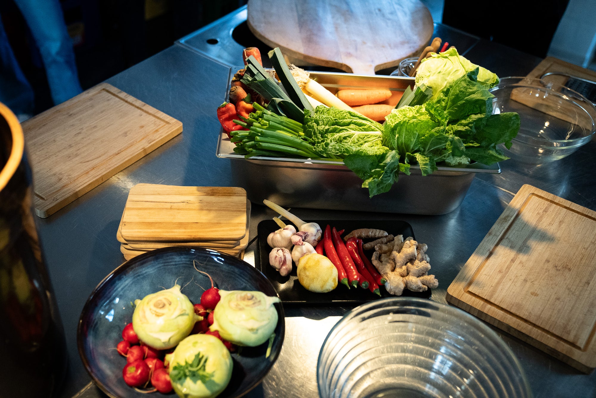 Events in Münster – Blick auf die Kochinsel in der Kochwerkstatt. Es sind Kohlrabi, Radieschen in einer Schüssel. Auf einem Teller liegt Inger, Chili und Knoblauch, wie auch Sellerie. In einem GN-Behälter sind Schnittlauch, Lauch, Möhren, Salat und Paprika. Drumherum stehen Schneidebretter und Lehreschüsseln, wie auch eine Reibe.