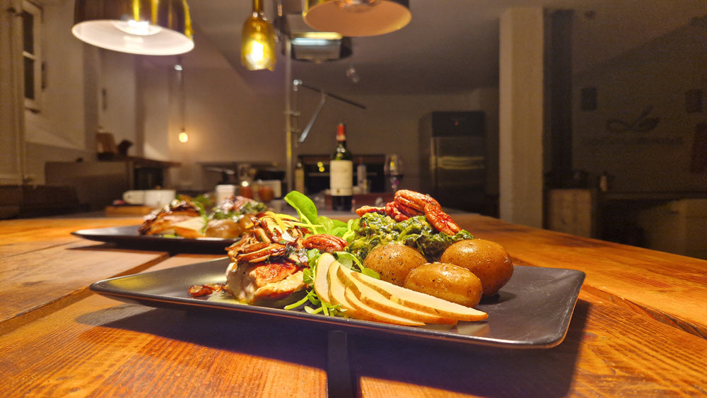 Teller mit Fleisch, Trockentomaten, Salzkartoffeln, Pesto und Salat. Im Hintergrund sieht man die Kochwerkstatt und einen weiteren Teller mit Essen, wie auch eine Rotweinflasche